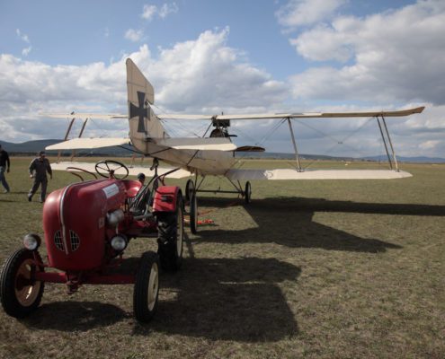 Ein historisches Flugzeug steht für die Flugvorbereitungen auf einer grüner Wiese bei strahlend blauem Himmel. Davor steht ein roter Traktor.