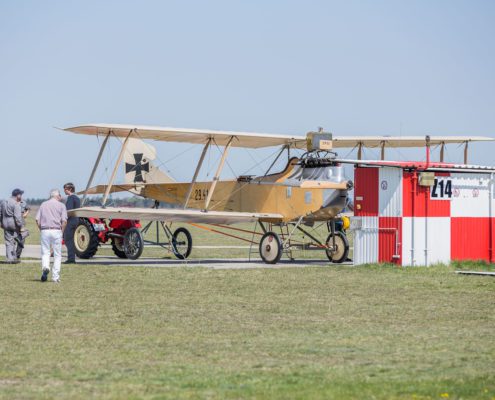 Historischer Doppeldecker wird von einem kleinen roten Traktor auf einem Flugfeld positioniert, umgeben von Helfern und Zuschauern.