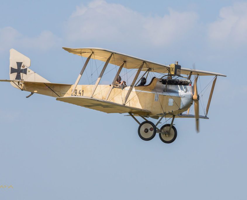 Ein zweisitziges Doppeldeckerflugzeug aus dem frühen 20. Jahrhundert fliegt bei strahlendem Sonnenschein. Die Markierungen deuten auf eine historische Militärmaschine hin